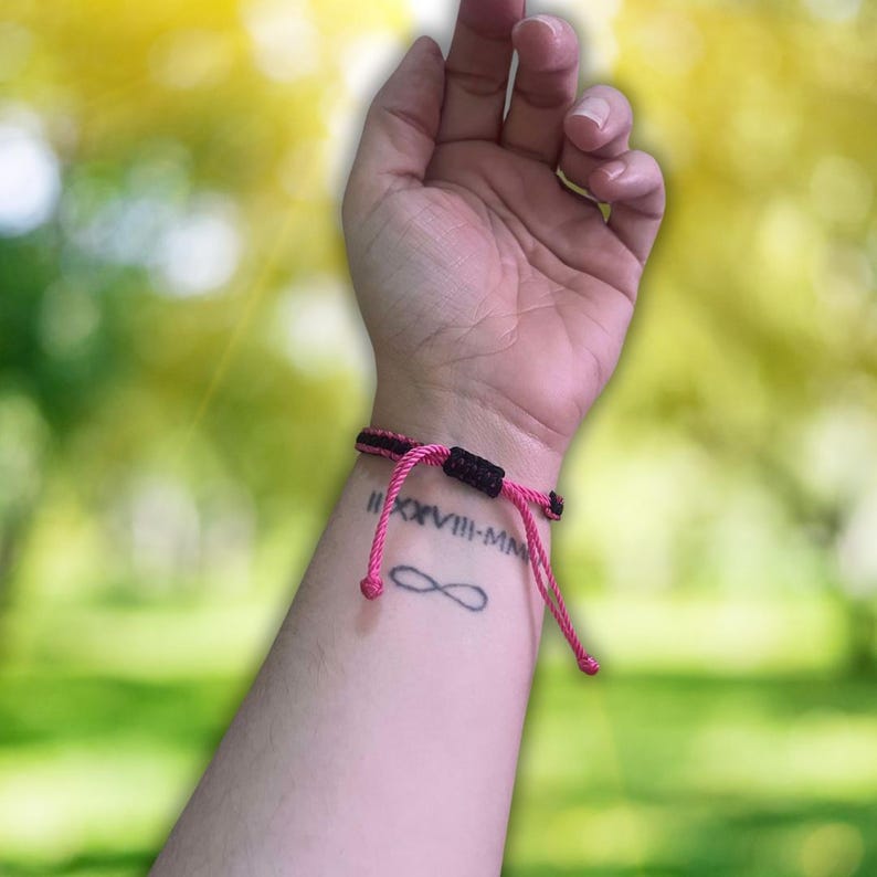 Pode incluir: Uma pulseira de corda rosa e preta com a inscri&ccedil;&atilde;o em algarismos romanos "II.XVIII-MM" e uma tatuagem de s&iacute;mbolo de infinito no pulso.