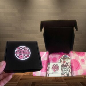 Pode incluir: Uma caixa preta com um logotipo rosa e preto que diz "Frankie's Fab Designs" e um padr&atilde;o xadrez rosa e preto. A caixa est&aacute; aberta e h&aacute; papel de seda rosa e branco dentro. O papel de seda tem um padr&atilde;o xadrez rosa e preto com um rosto sorridente rosa.