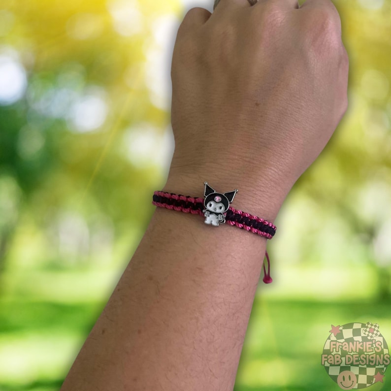 Pode incluir: Um bracelete de amizade preto e rosa com um amuleto prateado com um personagem de desenho animado com cabelo preto e um la&ccedil;o rosa. O amuleto tem uma &uacute;nica gema rosa no peito.