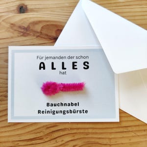 Op de afbeelding: Een wenskaart met Duitse tekst, waaronder "ALLES" en "Bauchnabel Reinigungsbürste", en een roze, pluizig object. Een witte envelop is gedeeltelijk zichtbaar. De kaart ligt op een houten oppervlak.