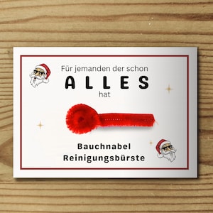 May include: A white rectangular card with a red border featuring a red pipe cleaner, two illustrations of a Santa Claus wearing sunglasses, and the German text: Für jemanden der schon ALLES hat Bauchnabel Reinigungsbürste.