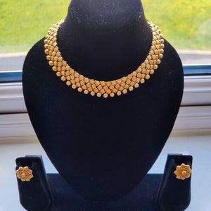 以下が含まれることがあります： 繊細なデザインのゴールドネックレスとマッチするイヤリング。ネックレスは、重なり合った葉のパターンで配置された小さな丸いビーズで作られています。イヤリングは、同様の葉のデザインのシンプルなスタッドです。