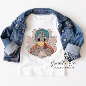 May include: White t-shirt with a cartoon turkey wearing a teal baseball cap. The turkey is brown with a red wattle and is holding its wings in front of it. The text "June & Co. DOODLES & DESIGNS" is at the bottom of the shirt.