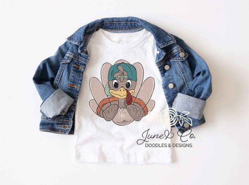 May include: White t-shirt with a cartoon turkey wearing a teal baseball cap. The turkey is brown with a red wattle and is holding its wings in front of it. The text "June & Co. DOODLES & DESIGNS" is at the bottom of the shirt.