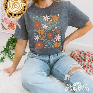 May include: A dark gray t-shirt with a floral design featuring orange, pink, and white flowers on a green leafy background. The text "Thank you for supporting my original art!" is on a yellow circle sticker.