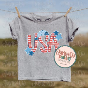 Op de afbeelding: Een grijs T-shirt met een rood-wit-blauwe USA-afbeelding. De afbeelding bevat een baseballpet, sterren en strepen. De tekst "June & Co. Dolls Original Art" staat in een roze cirkel met een witte rand.