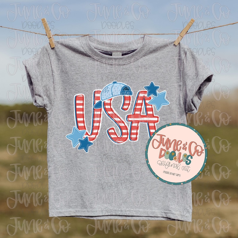 Op de afbeelding: Een grijs T-shirt met een rood-wit-blauwe USA-afbeelding. De afbeelding bevat een baseballpet, sterren en strepen. De tekst "June & Co. Dolls Original Art" staat in een roze cirkel met een witte rand.