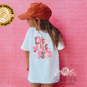 May include: A white t-shirt with the text "Love Like Jesus" in pink and red letters with hearts. The shirt is being worn by a child wearing a brown baseball cap.
