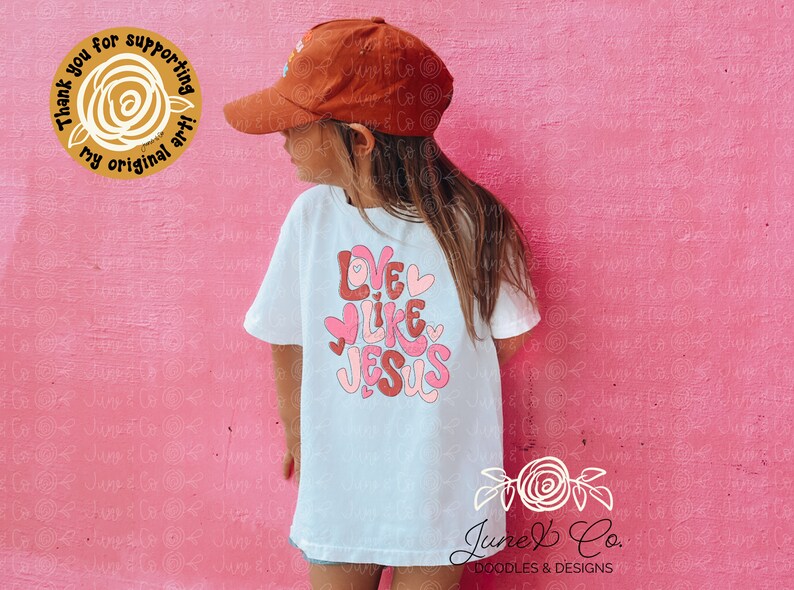 May include: A white t-shirt with the text "Love Like Jesus" in pink and red letters with hearts. The shirt is being worn by a child wearing a brown baseball cap.