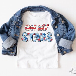 Puede incluir: Camiseta blanca con el texto "Oh My Stars" en rojo, blanco y azul con un patr&oacute;n de bandera estadounidense desgastado.