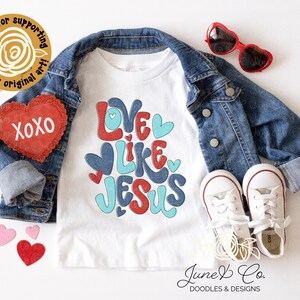 Peut inclure: T-shirt blanc pour enfant avec le texte "Love Like Jesus" en lettres rouges, bleues et turquoise avec des c&oelig;urs. Le t-shirt est pr&eacute;sent&eacute; sur un fond blanc avec une veste en jean, des lunettes de soleil en forme de c&oelig;ur rouges et des c&oelig;urs en feutre rouge.