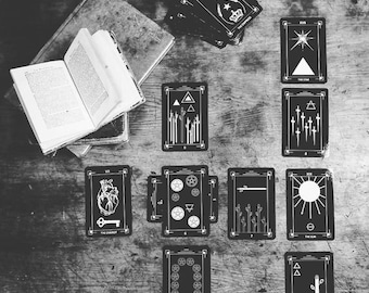 May include: Black and white tarot cards with various symbols and illustrations. The cards are laid out on a wooden surface. The cards include The Chariot, The Star, The Sun, and The Knight.