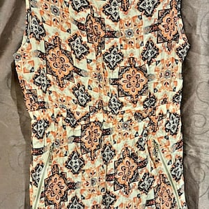 May include: A sleeveless white dress with a geometric pattern in shades of orange, black, and grey. The dress has a drawstring waist and side pockets.