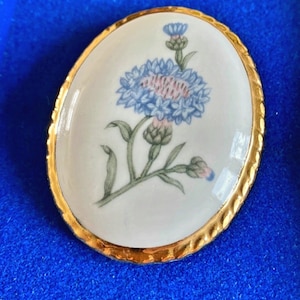 Op de afbeelding: Een ovale broche van wit porselein met een gouden rand, met een blauwe en roze bloem met groene bladeren.