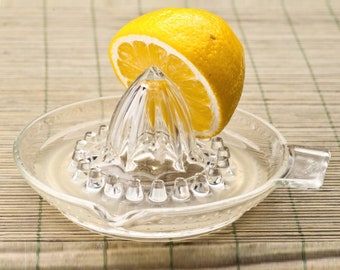 May include: A clear glass citrus juicer featuring a central ridged cone for extracting juice and a circular basin with a small handle and a pouring spout. A bright yellow half of a fresh lemon is placed directly onto the juicer's central cone.
