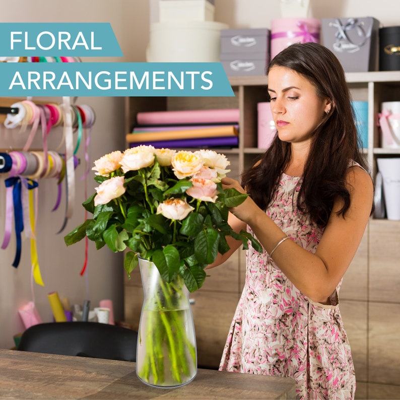 May include: A woman arranges a bouquet of light pink and cream roses in a clear glass vase. The vase has a textured surface. The sign above reads "Floral Arrangements".