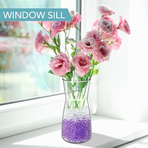 May include: A clear glass vase with a wide mouth and a narrow base filled with purple and clear water beads. Pink flowers are arranged in the vase. The vase is sitting on a white window sill.