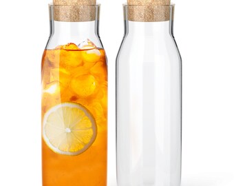 Può includere: Due caraffe in vetro trasparente con corpo cilindrico e collo affusolato, ciascuna dotata di un tappo in sughero naturale. Una caraffa è riempita con un liquido arancione, cubetti di ghiaccio e una fetta di limone giallo che galleggia.
