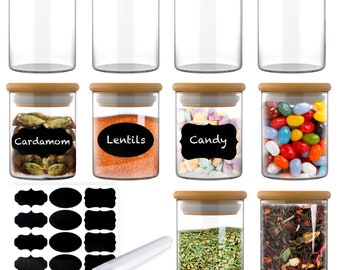 Può includere: Un set di barattoli in vetro trasparente con coperchi in legno marrone chiaro. Alcuni barattoli contengono cardamomo, lenticchie, caramelle, erbe essiccate e foglie di tè, con etichette nere e testo bianco. Include un foglio di etichette vuote.