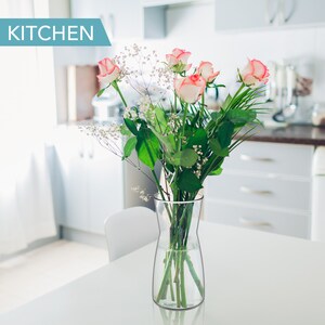 May include: A bouquet of pink and white roses with green leaves in a clear glass vase on a white table in a kitchen.