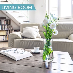May include: A white couch with a white throw pillow in a living room with a wooden coffee table. A clear glass vase with white flowers and green leaves sits on the table. A book and a pair of glasses are also on the table. A cup of coffee sits on a saucer next to the vase.