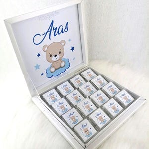 Könnte beinhalten: Eine weiße Geschenkbox mit einem Deckel, der sich öffnet und ein Tablett mit einzeln verpackten Pralinen enthüllt. Der Deckel hat ein blaues und weißes Design mit einem Comic-Bären und dem Namen "Aras" in blau. Jede Praline ist in durchsichtige Folie mit einem blauen und weißen Design verpackt und der Name "Aras" ist darauf gedruckt.