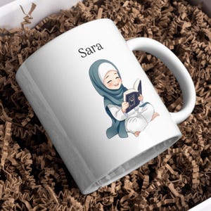 Könnte beinhalten: Ein weißer Keramikbecher mit einer Cartoon-Illustration einer lächelnden Person, die einen blaugrünen Hijab und weiße Kleidung trägt und sitzend ein offenes Buch liest. Der Name Sara ist in schwarzer Schrift über der Illustration aufgedruckt.