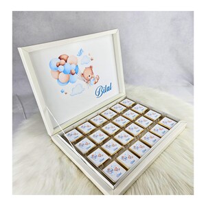 Könnte beinhalten: Eine weiße Box mit einem Deckel, der sich öffnet und ein Tablett mit kleinen Schokoladenquadraten enthüllt. Der Deckel zeigt eine Aquarell-Illustration eines Teddybären, der Ballons hält, und den Namen "Bilal".