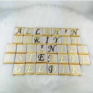 Könnte beinhalten: Anordnung rechteckiger, goldumrandeter, weißer Kacheln mit goldener und schwarzer Schrift. Die Kacheln buchstabieren eine Nachricht auf Türkisch. Die goldene Schrift ist in einer Schreibschrift. Die Kacheln sind auf einer weißen, flauschigen Oberfläche angeordnet.