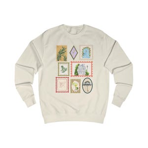 May include: A cream-colored crewneck sweatshirt featuring a graphic print of various postage stamp designs. The stamps display illustrations of a green plant, a blue door, a frog, a house, a cocktail, and a black umbrella on a light background.