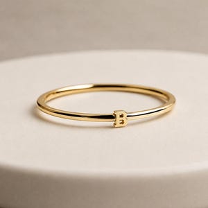 Puede incluir: Un anillo fino de tono dorado pulido que presenta una única letra mayúscula B pequeña centrada en la banda. El anillo tiene un perfil liso y redondeado con un acabado metálico reflectante, mostrando un diseño sencillo y limpio sin adornos adicionales.