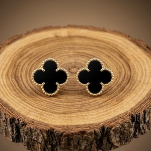 May include: A pair of stud earrings featuring a four-leaf clover shape with a smooth black center inlay and a gold-toned beaded border. The earrings have a symmetrical design with a polished finish, showcasing a classic floral-inspired silhouette.