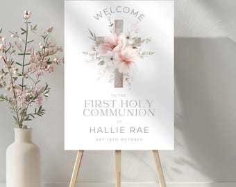 May include: Welcome to the First Holy Communion of Hallie Rae, Sat 18th October. A white sign with a watercolor illustration of a wooden cross with pink flowers and greenery. The sign is on a wooden easel.