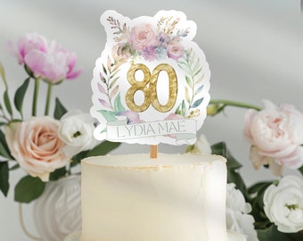 Könnte beinhalten: Ein weißer Kuchen mit einem floralen Kuchentopper mit der Zahl 80 in Gold und dem Namen "LYDIA MAE" auf einem Banner.