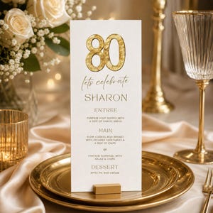 May include: A gold-colored menu card for an 80th birthday celebration. The card reads "Let's Celebrate Sharon" and lists the menu: pumpkin soup, slow cooked beef brisket, or chicken schnitzel, and apple pie. The card is on a gold plate.