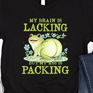 Könnte beinhalten: Schwarzes T-Shirt mit einem Cartoon-Frosch und dem Text "MY BRAIN IS LACKING BUT MY ASS IS PACKING" in Grün und Gelb. Der Frosch ist hellgrün und lächelt, umgeben von blauen Blumen.