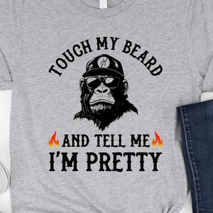 Puede incluir: Camiseta gris jaspeado con un diseño de un gorila con gafas de sol y gorra. El texto en la camiseta dice "TOUCH MY BEARD AND TELL ME I'M PRETTY" en letras negras con llamas a ambos lados.