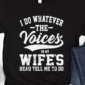 Może przedstawiać: Czarny t-shirt z białym napisem: "I DO WHATEVER -THE- Voices IN MY WIFE'S HEAD TELL ME TO DO." Koszulka jest złożona na dole. Widoczne są białe trampki i niebieskie dżinsy.