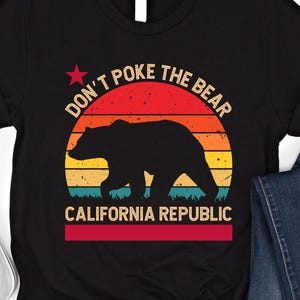 May include: Black t-shirt featuring a graphic of a bear silhouette walking in front of a sunset design. The text "DON'T POKE THE BEAR" arches above the bear, with "CALIFORNIA REPUBLIC" below. A red star is above the text.
