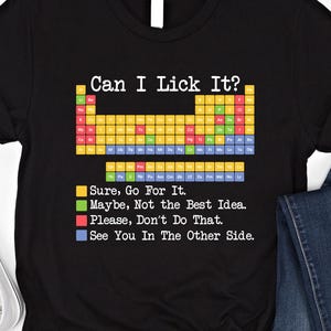 以下が含まれることがあります： 「Can I Lick It?」の文字とカラフルな周期表のデザインがプリントされた黒いTシャツ。その下に、色分けされたフレーズがあります。「Sure, Go For It」「Maybe, Not the Best Idea」「Please, Don't Do That」「See You In The Other Side」.