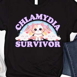 Puede incluir: Camiseta negra con un arcoíris y un gato de dibujos animados. El texto "Chlamydia Survivor" está escrito en letras moradas.