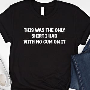 Peut inclure: T-shirt noir avec texte blanc : "THIS WAS THE ONLY SHIRT I HAD WITH NO CUM ON IT." Le t-shirt a un col rond. Des baskets blanches et un jean bleu sont partiellement visibles en arrière-plan.