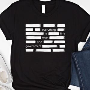 Puede incluir: Camiseta negra con texto blanco: "everything is fine, trust your government". El texto está dispuesto en un bloque rectangular con líneas horizontales. La camiseta se combina con vaqueros azules y zapatillas blancas.