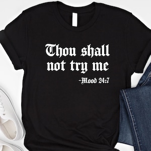 Puede incluir: Camiseta negra con el texto blanco "Thou shall not try me - Mood 24:7". La camiseta tiene mangas enrolladas y un nudo en la parte inferior. Zapatillas blancas y vaqueros azules son visibles en el fondo.