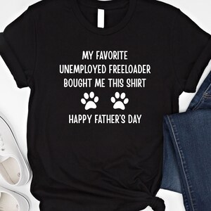 May include: Black t-shirt with white text: "MY FAVORITE UNEMPLOYED FREELOADER BOUGHT ME THIS SHIRT" above two paw prints and "HAPPY FATHER'S DAY". The shirt is styled with rolled sleeves and a knot at the bottom.