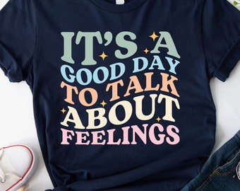 Op de afbeelding: Een marineblauw t-shirt met de tekst "It's a good day to talk about feelings" in kleurrijke retro-lettertypen.