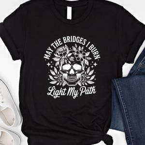 May include: Black t-shirt with a white skull graphic surrounded by flowers and leaves. The text "MAY THE BRIDGES I BURN" arches above the skull, with "Light My Path" below. The shirt is paired with blue jeans and white sneakers.