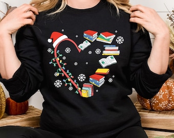 May include: Black sweatshirt with a heart-shaped design featuring a Santa hat, string lights, snowflakes, and stacks of books. The design is in red, white, green, and yellow. The sweatshirt is a long-sleeved crew neck.