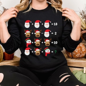 以下が含まれることがあります： クリスマスをテーマにした数学の方程式のデザインが施された、黒色の長袖スウェットシャツ。 方程式には、サンタ、トナカイ、雪だるまのグラフィックと、加算、減算、乗算の記号が含まれています。 最後の式の答えは不明です。