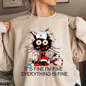 Może przedstawiać: Beżowa bluza z czarnym kotem z dużymi oczami, w czepku pielęgniarki i czapce Świętego Mikołaja. Kot jest opleciony kolorowymi lampkami choinkowymi, ze stetoskopem i napisem "IT'S FINE. I'M FINE. EVERYTHING IS FINE."